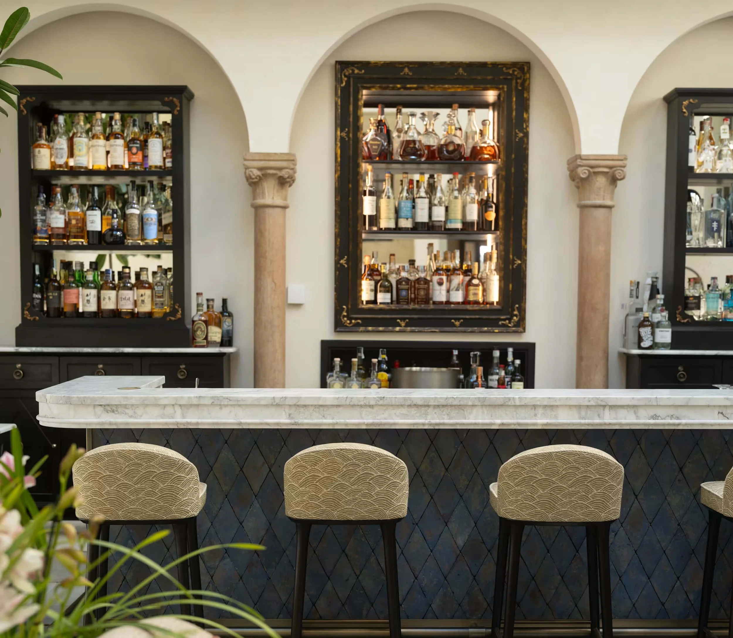 Bar counter with elegant chairs and black shelves display bottles of liquor in a stylish interior with arches and columns.