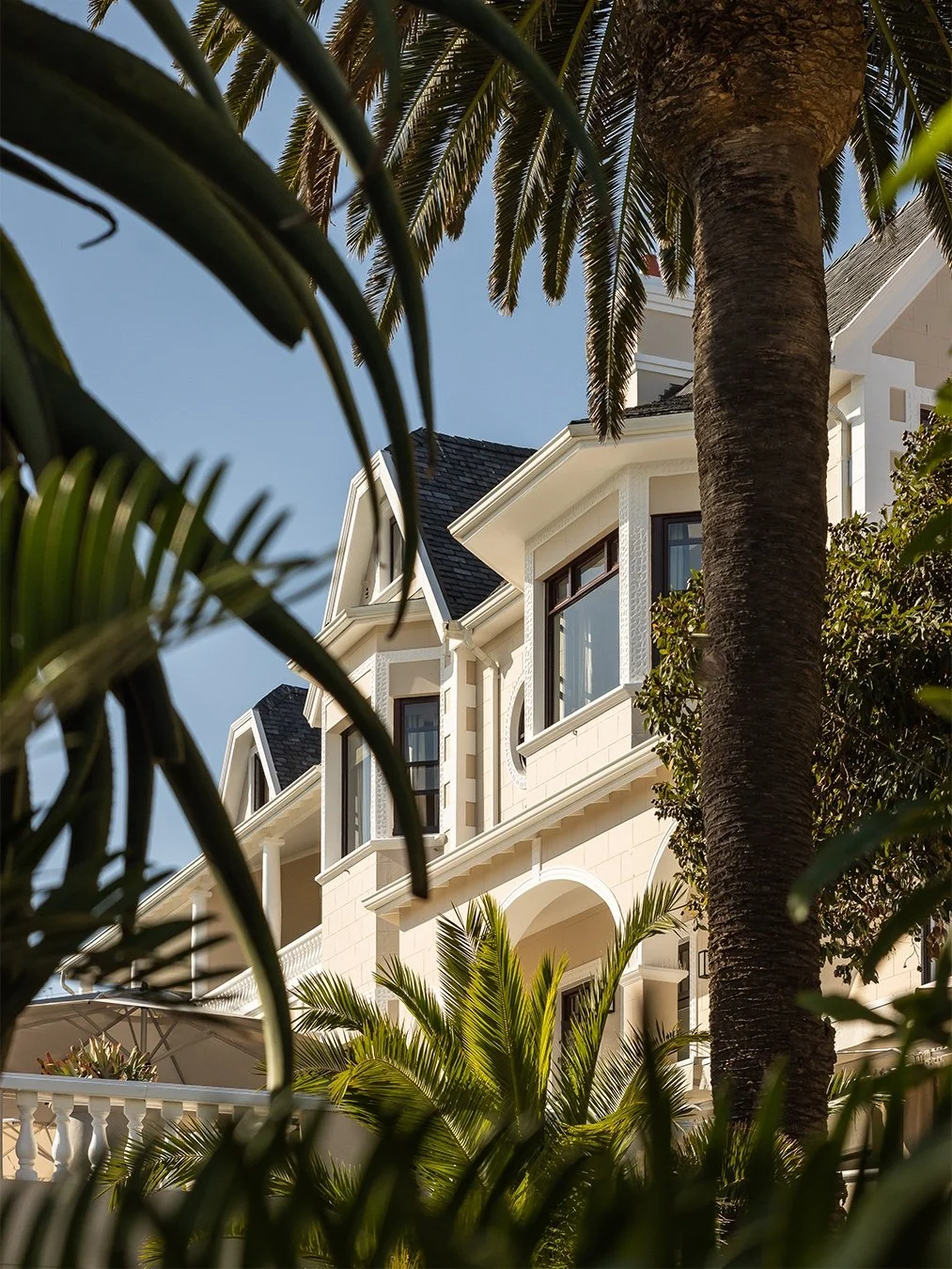 Partially visible multi-story house with white exterior, large windows, black roof, surrounded by tropical palm trees and greenery.