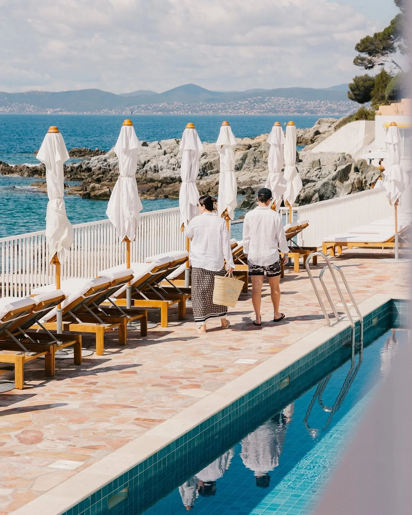 Two people walking along a poolside area with lounge chairs and closed umbrellas, overlooking rocky coastline and water with hills in the distance.