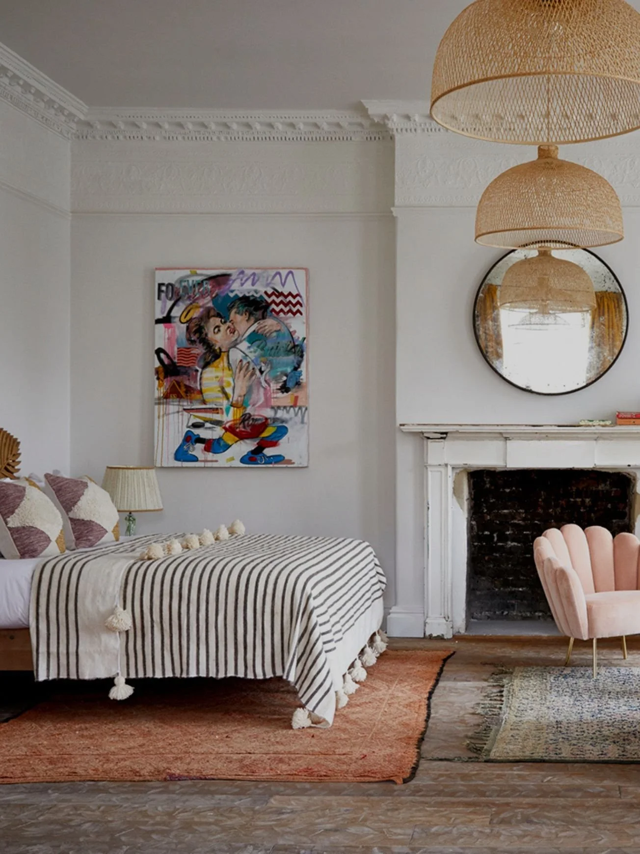 A cozy living room corner with a striped beige and white sofa, a table with pom-pom trim, and a pink velvet chair. Wall art featuring a colorful pop art piece, a round mirror above the fireplace, and woven hanging lamps provide warm lighting. The room has a white fireplace mantle, a textured rug, and vintage-style decor.