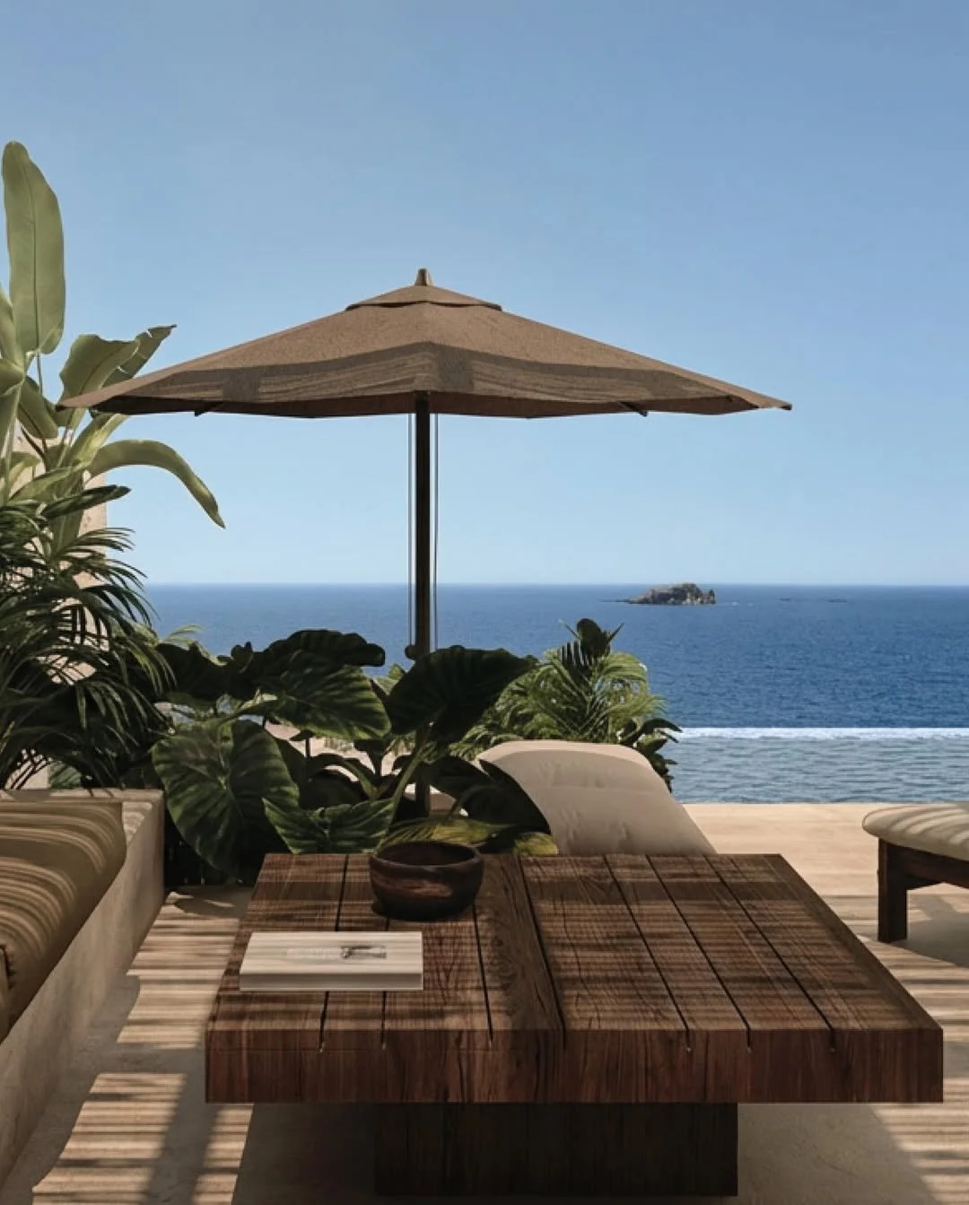 A wooden outdoor dining table with a book and a small bowl, surrounded by lounge chairs, with an umbrella, overlooking the ocean and a small island in the distance.
