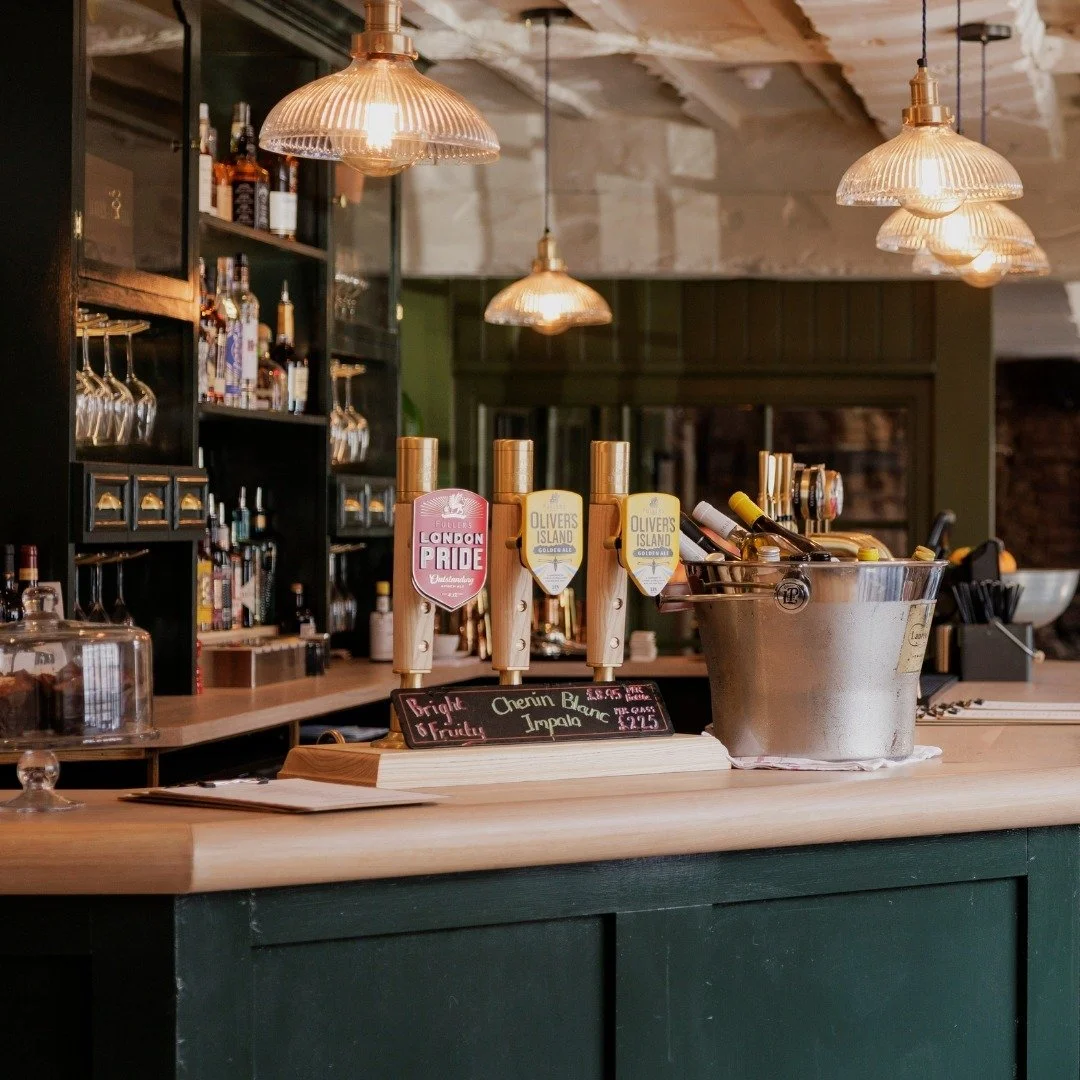 A bar counter with beer taps, a bucket of wine bottles, and hanging pendant lights in a cozy, rustic-style bar or cafe.