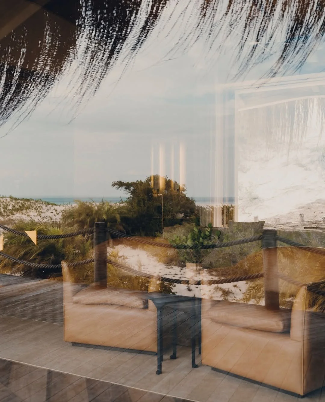 Reflection of a beach scene with sand dunes, greenery, and a cloudy sky seen through a window from inside a room, showing chairs and a table.