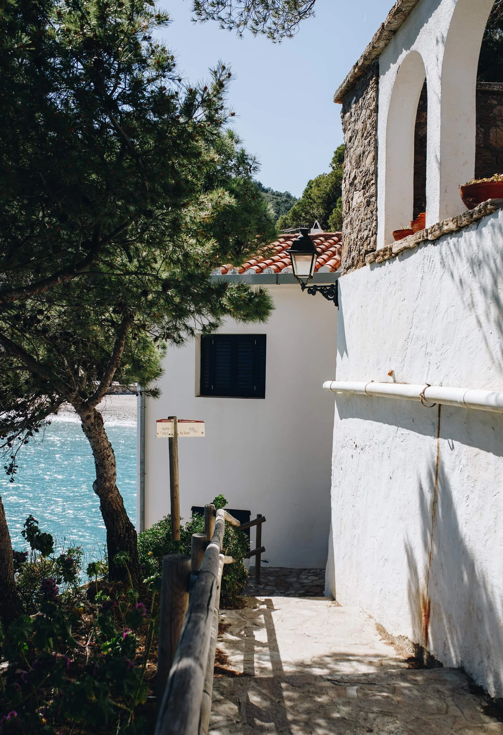 White stucco building with arched window frames and a stone wall, overlooking the sea, with a tree and a narrow walkway with a wooden railing and potted plants.