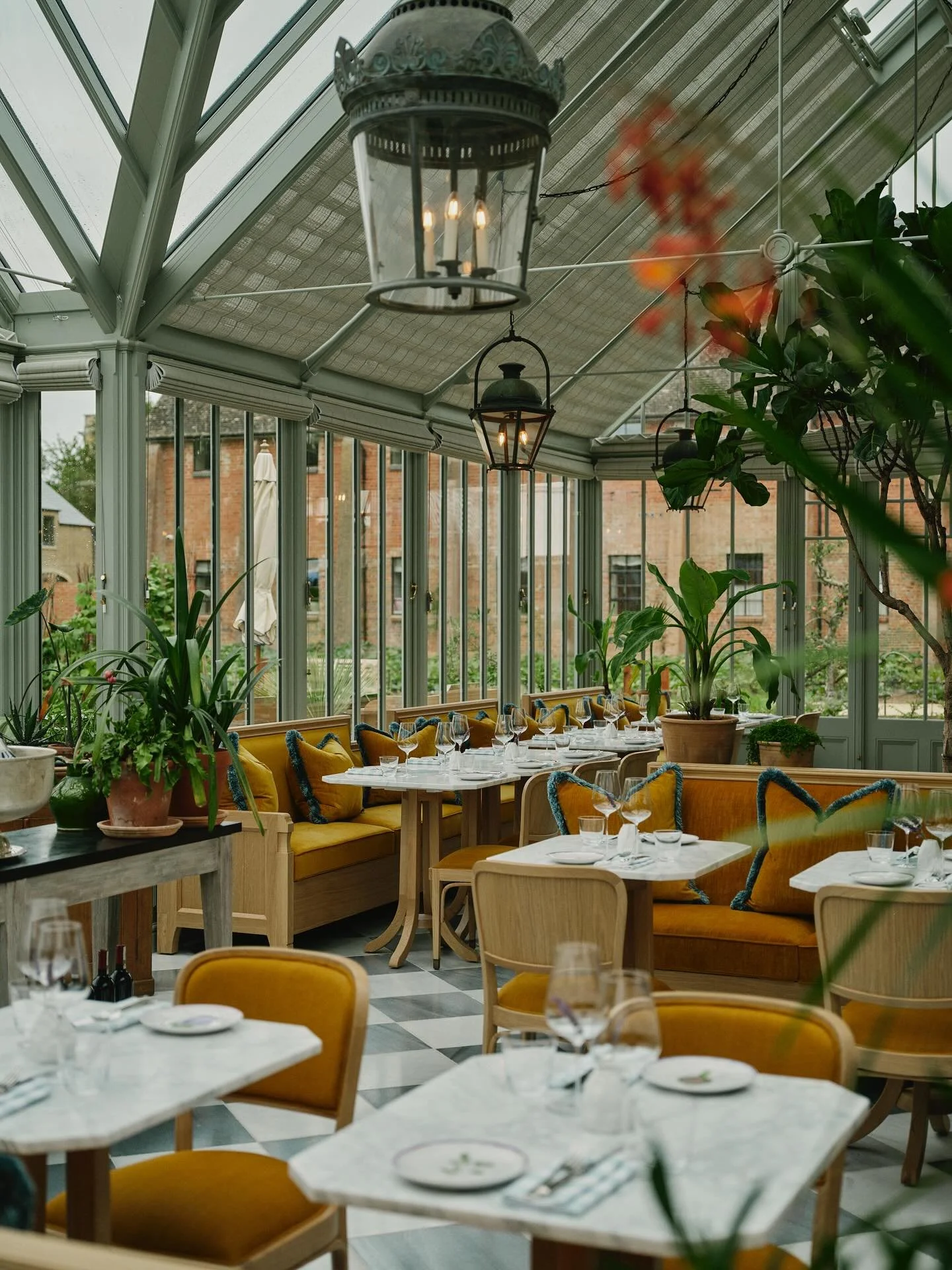 Interior view of a bright, conservatory-style restaurant with large glass windows, hanging lanterns, and lush green plants, set for dining with white tables, yellow upholstered chairs, and place settings.