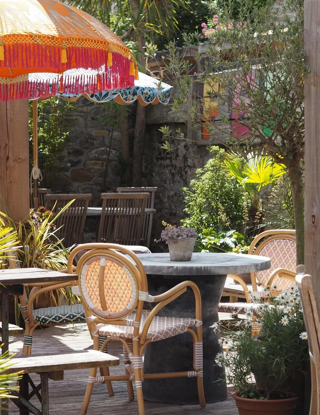 Outdoor patio with wooden furniture, a marble table, potted plants, garden chairs, colorful parasols, and lush greenery.