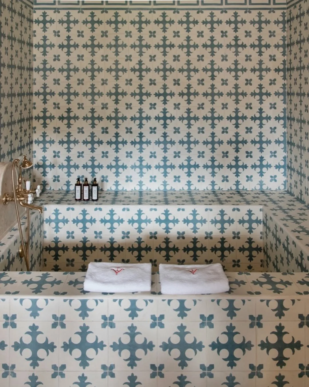 An in-wall bathtub lined with blue and white patterned tiles, with two folded white towels on the edge, a vintage brass bathtub faucet, and three bottles of bath products on the side.