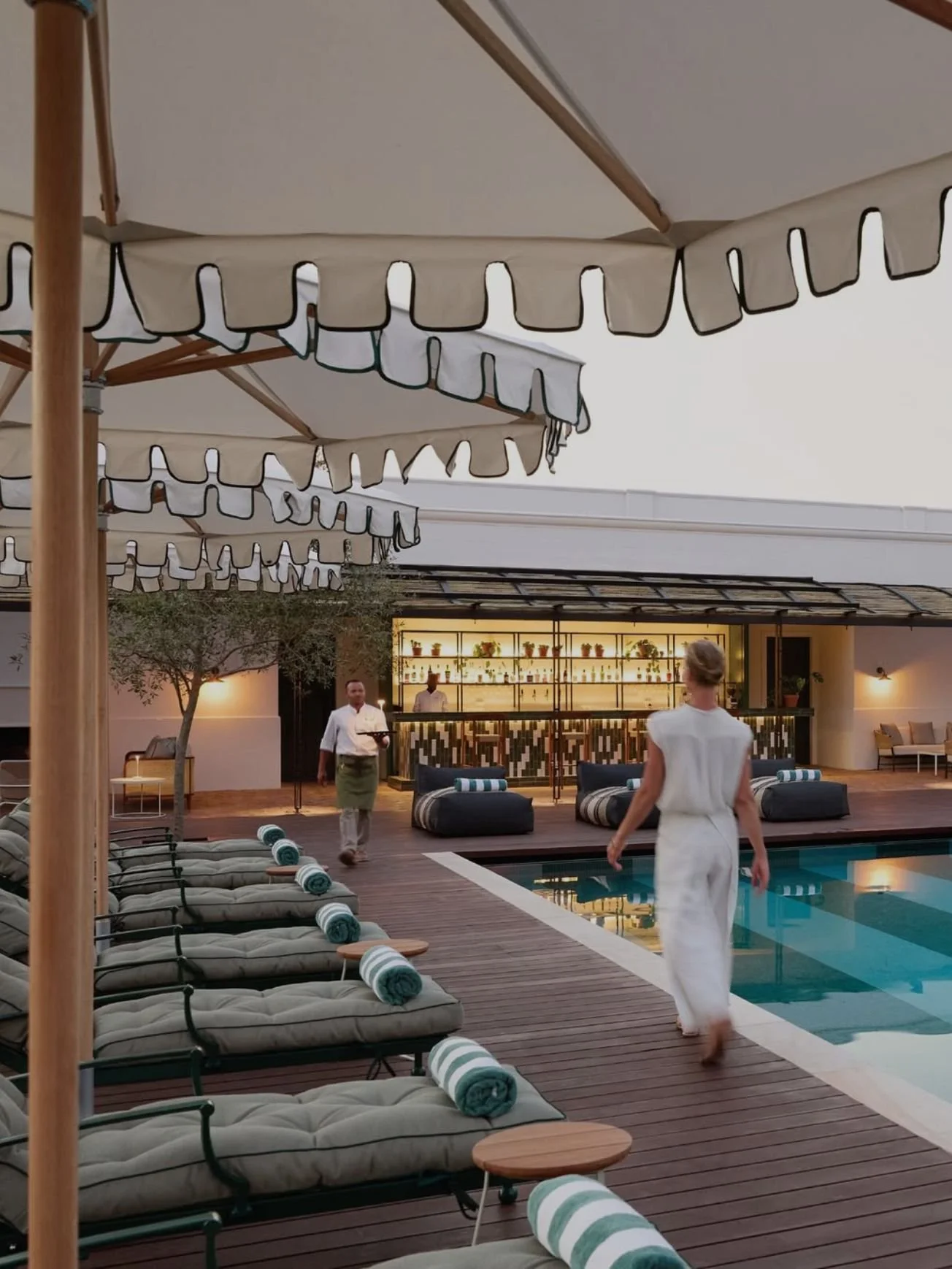 Poolside area with lounge chairs and umbrellas, a bar area in the background, and people walking around.