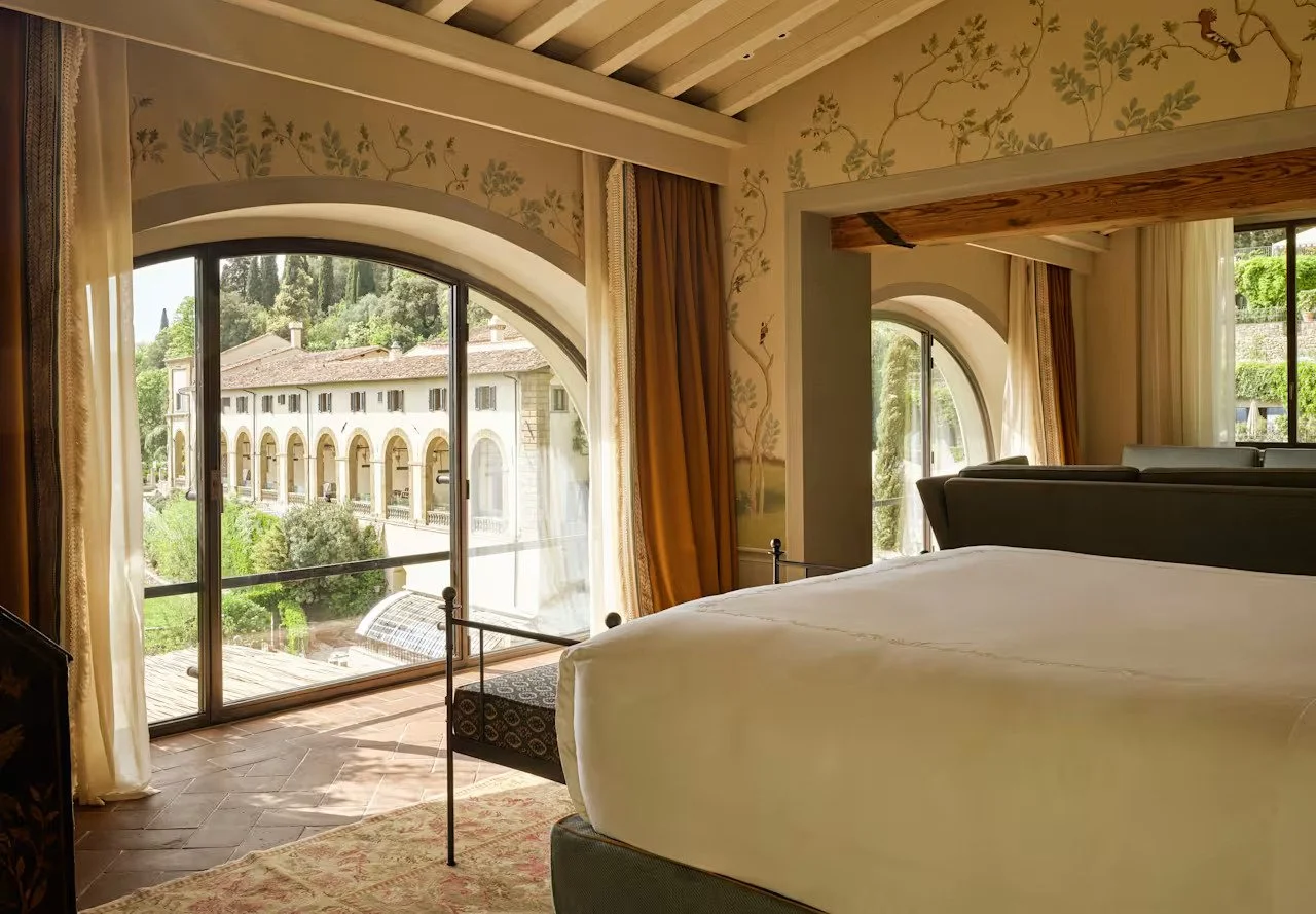 A bedroom with large arched windows showing a view of a historic building and greenery outside. The room has floral wallpaper, curtains, a bed with white bedding, and a tiled floor.