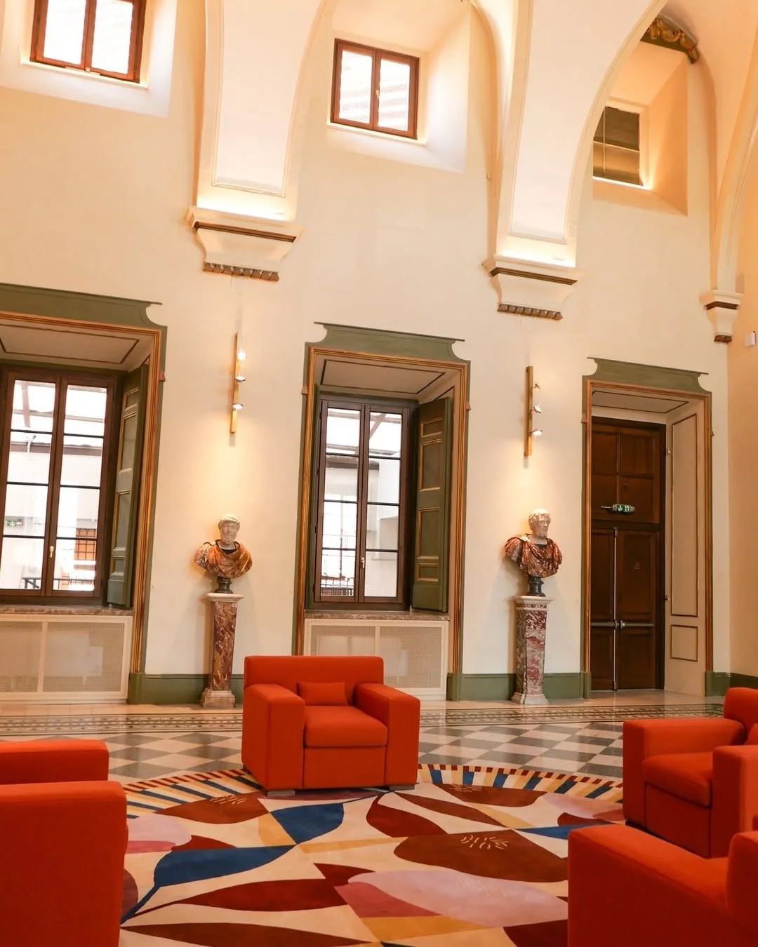 Elegant room with high arched ceilings, large windows, classical bust sculptures on pedestals, vibrant orange armchairs, and a geometric patterned carpet.
