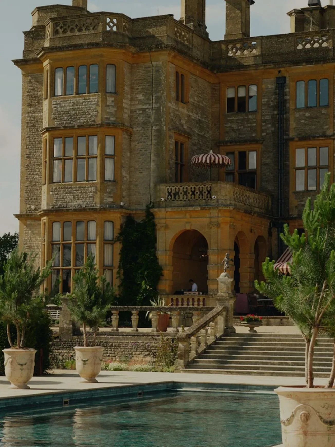 A large, historic stone building with multiple windows, balconies, and decorative elements. In front, there are stairs leading up to an arched entrance, surrounded by greenery and potted plants. A swimming pool is visible at the bottom of the image.