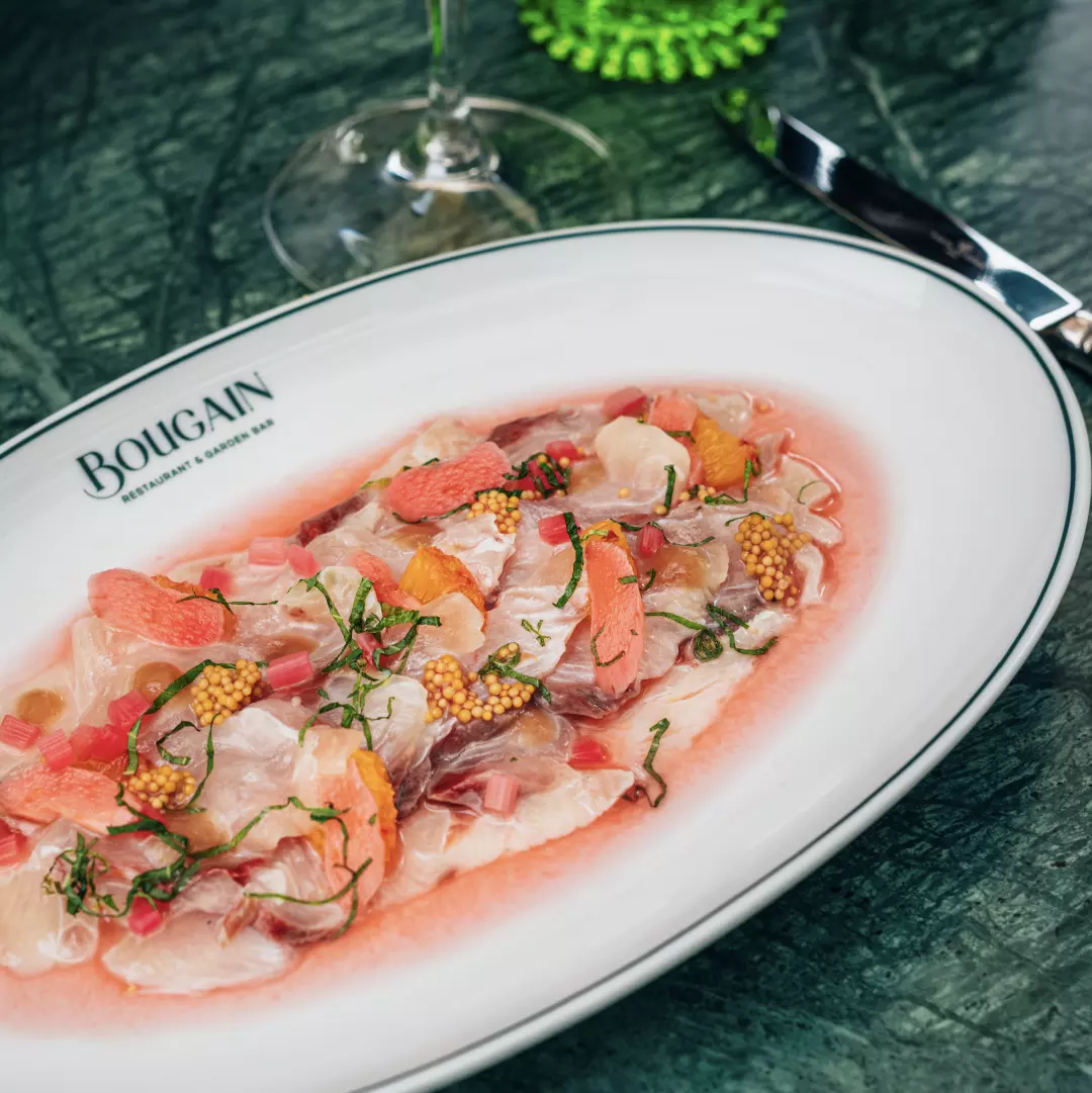 A plate of ceviche with raw fish, orange slices, herbs, and mustard seeds, on a green marble table with a glass of white wine and a black-handled utensil.