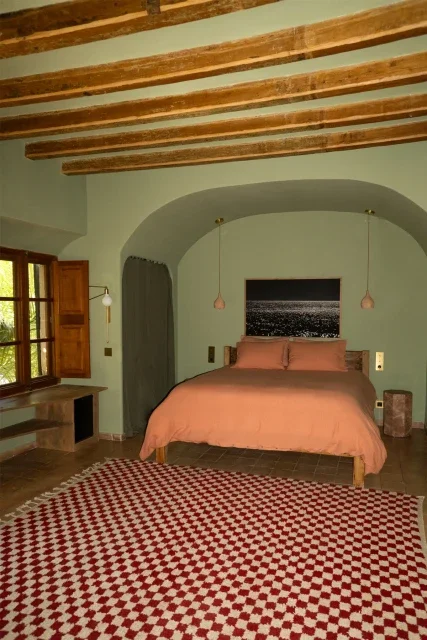 A bedroom with a green wall, wooden beams on the ceiling, a bed with peach-colored bedding, and a black and white cityscape picture above the bed. There are two pink hanging pendant lights, a window with wooden shutters, and a red and white patterned rug.
