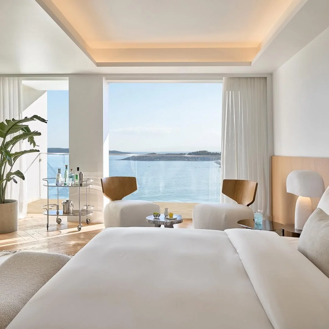 A modern hotel room with a large window overlooking a body of water and distant land, furnished with a bed, chairs, a bedside table, a lamp, a small round table with drinks, and a tall plant.