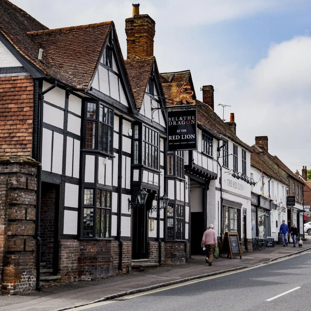 A street view of a traditional English pub with black and white timber framing, red brick accents, and a sign that reads 'Bell & The Dragon at the Red Lion.' People walk along the sidewalk, and the pub features decorative lanterns and a chalkboard sign at the entrance.
