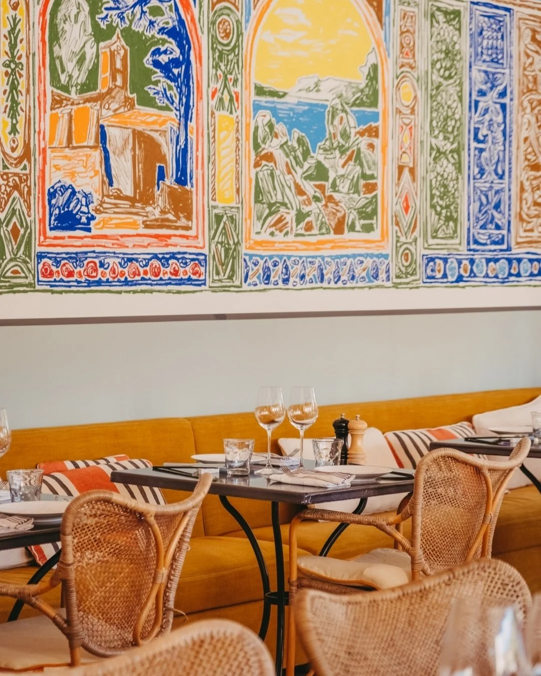 Colorful mural featuring scenic landscapes and architectural elements, seen above a restaurant with tables, glassware, and rattan chairs.