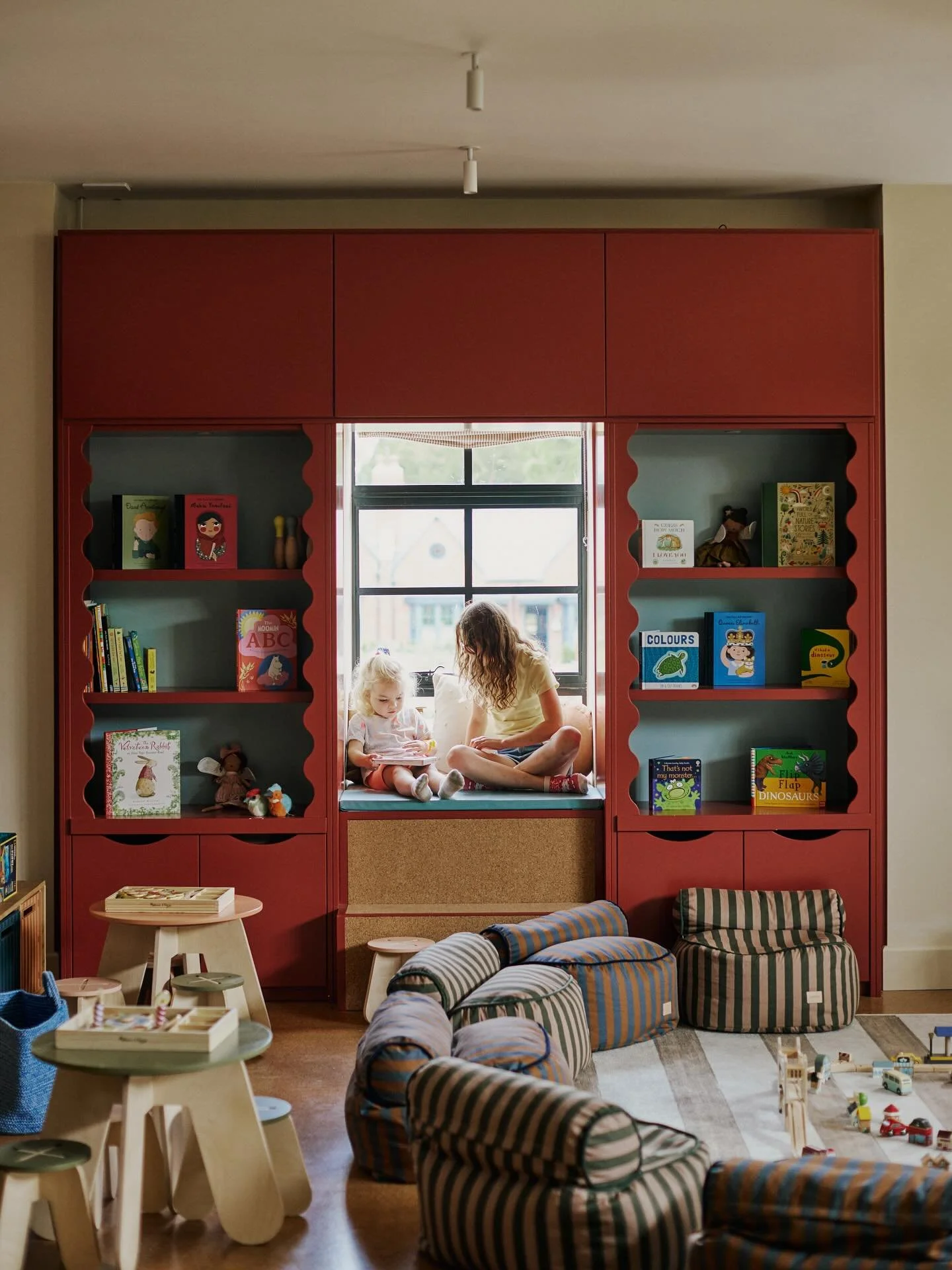 A cozy playroom with a red bookshelf next to a window seat where a woman and a child are sitting. The bookshelf contains children's books, toys, and stuffed animals. In front, there are striped bean bag chairs, a small wooden table with toys, and a play area with toy vehicles on the floor.