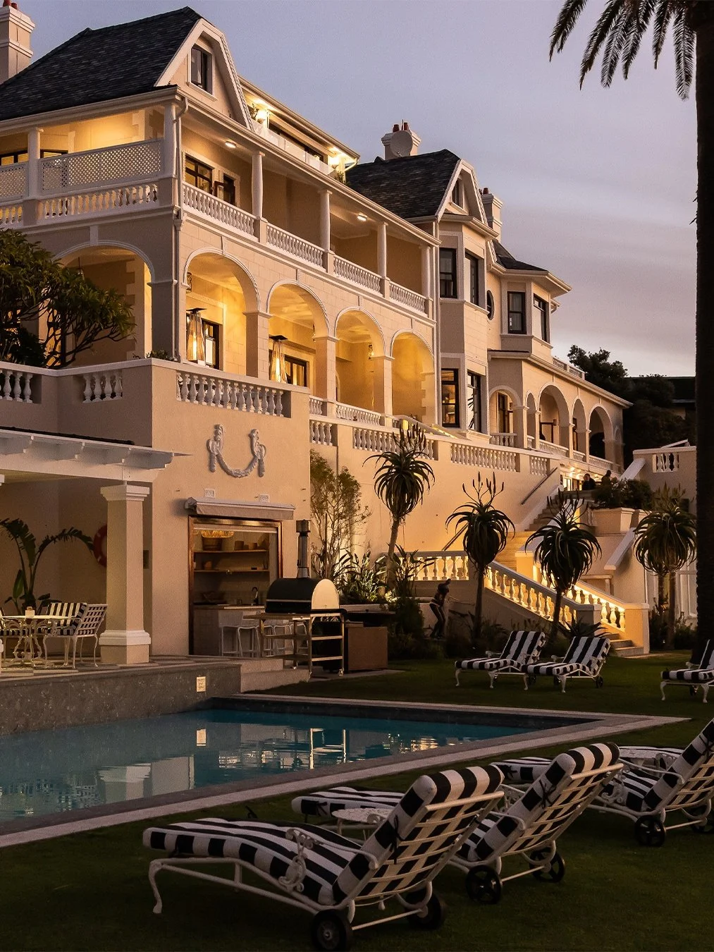 Luxury mansion with multiple balconies, exterior lighting, a swimming pool, and poolside lounge chairs at sunset