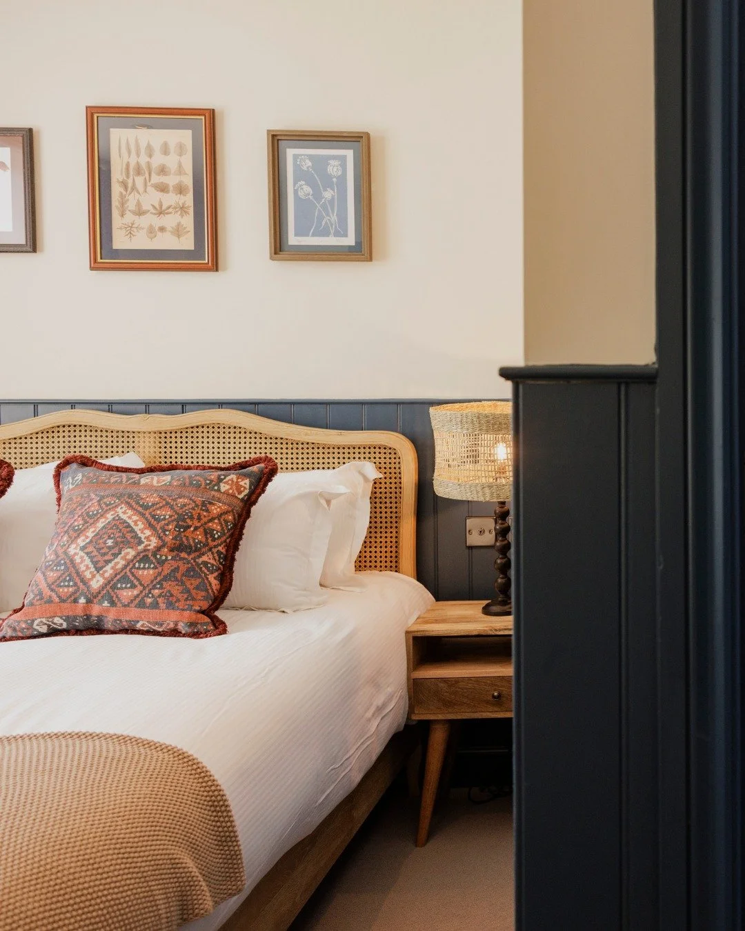 A cozy bedroom with a bed that has a wooden headboard and white bedding. There is a decorative pillow with a red and black geometric pattern. A nightstand holds a lamp with a woven shade, and framed botanical artwork hangs on the wall above the bed.