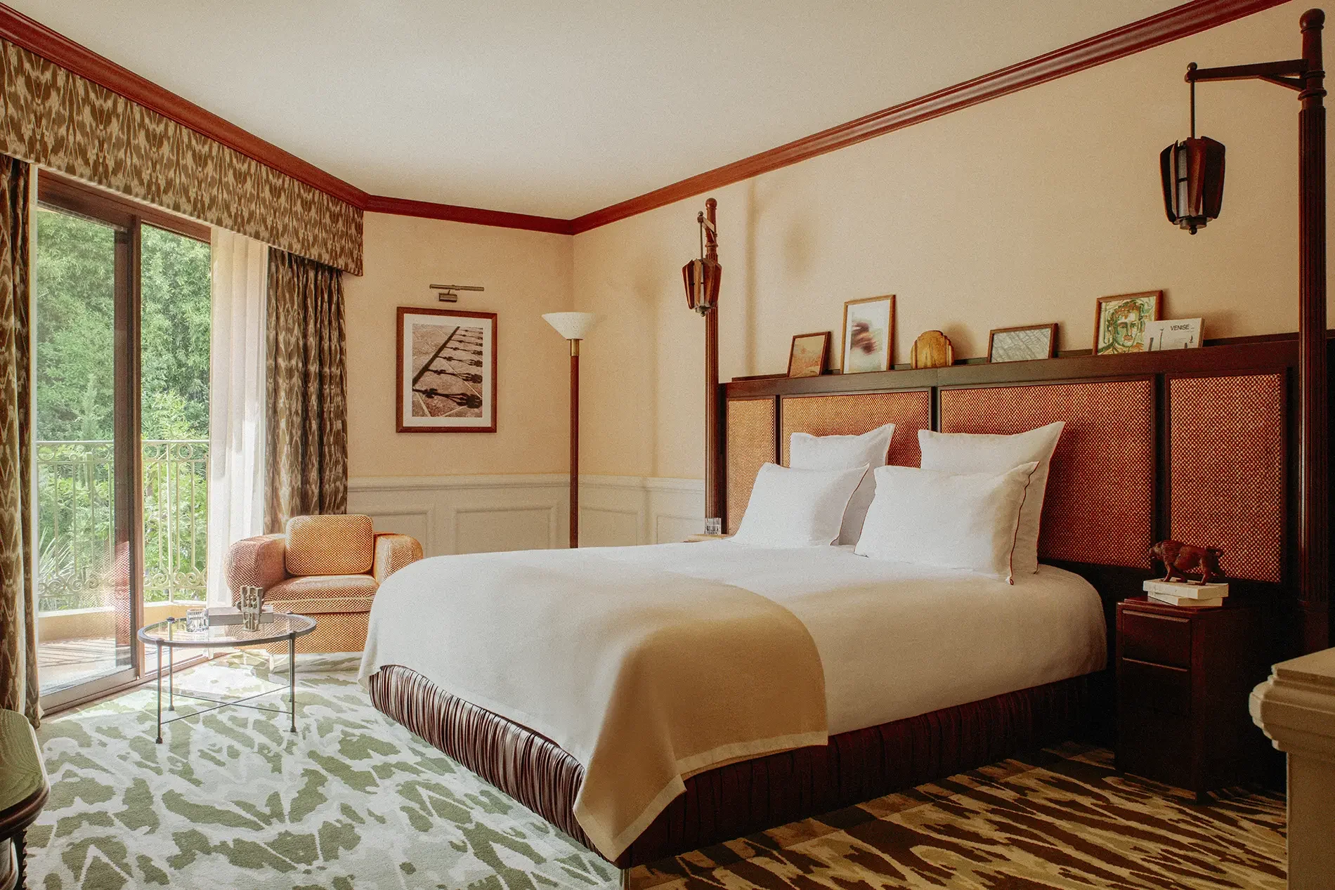 A hotel room with a large bed, decorative headboard, framed artwork, floor lamp, an orange armchair, glass door opening to a balcony with greenery outside, patterned curtains, and a green and white patterned area rug.