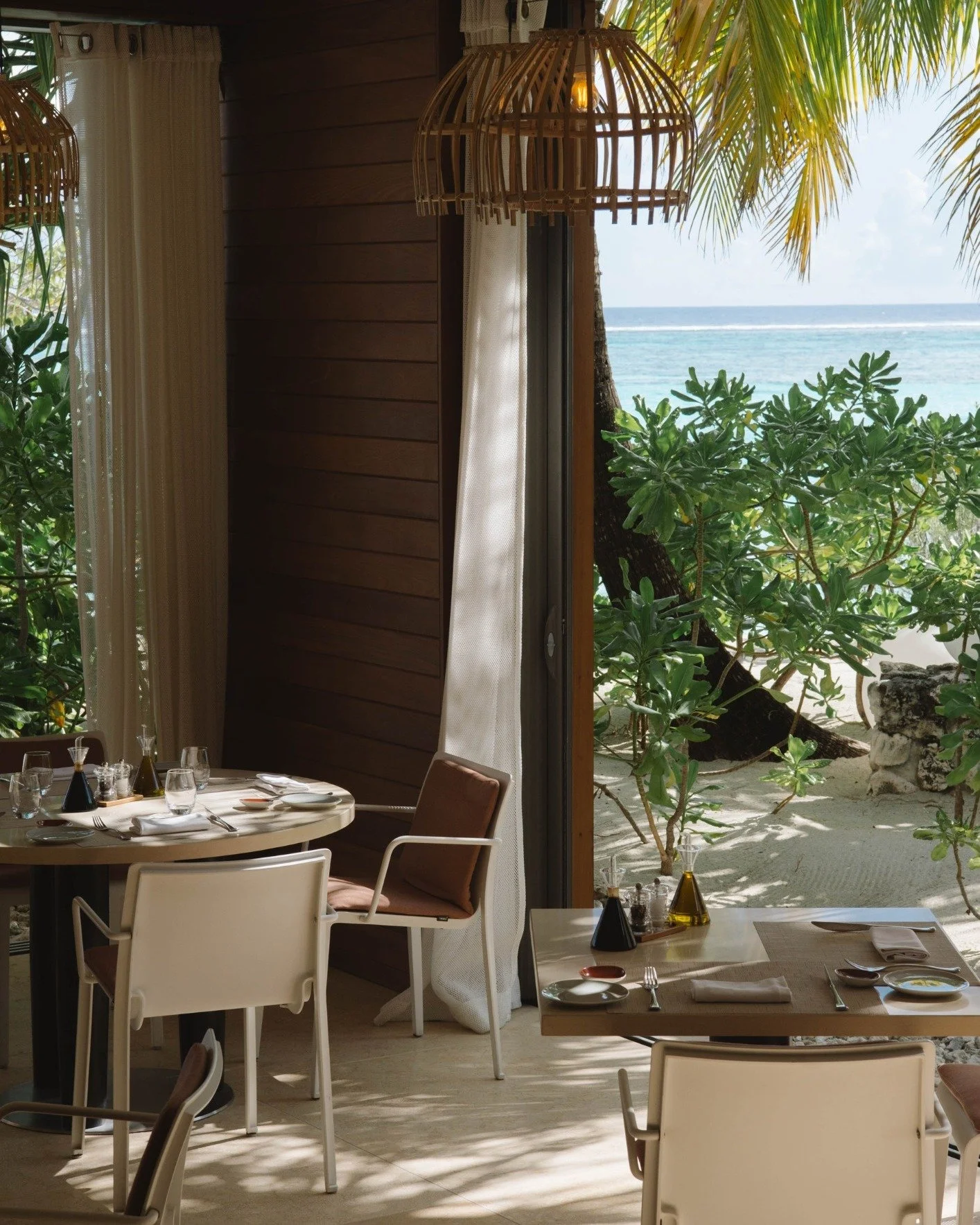A view of an indoor dining area with a table set for a meal, overlooking a tropical beach through an open door. The table has plates, glasses, and condiments, and the decor includes hanging wicker lamps. Outside are green plants, sandy ground, and the ocean in the background.