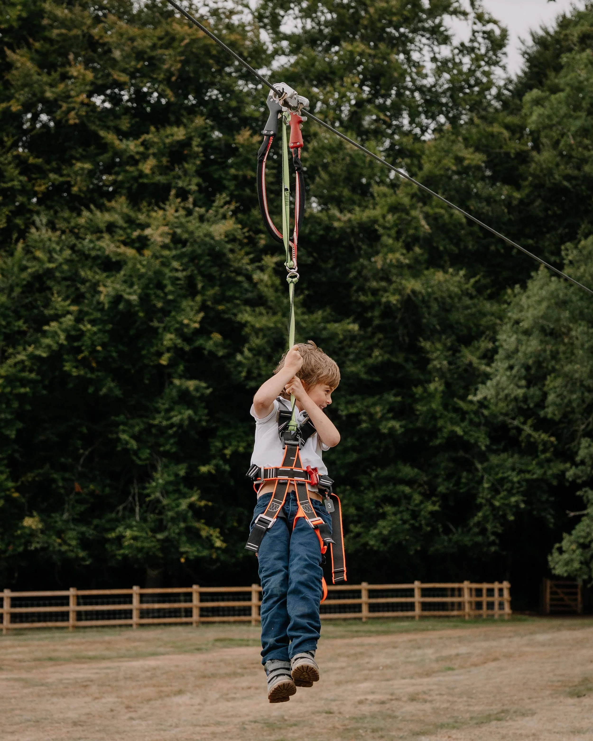A young child wearing a harness and holding onto a zip line handle, flying through the air outdoors with trees in the background.