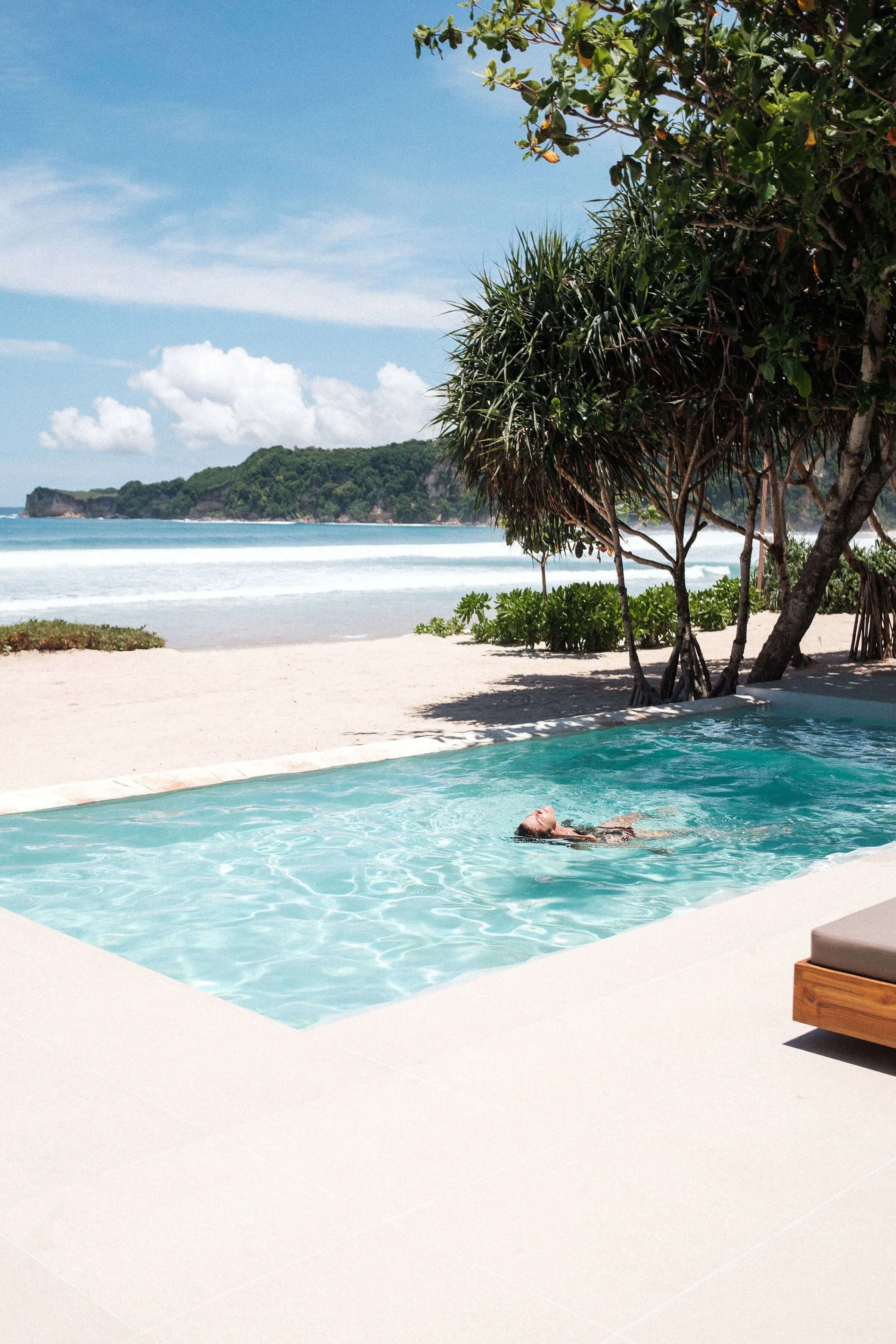 A woman relaxing and floating in a swimming pool near a tropical beach with trees and hills in the background.