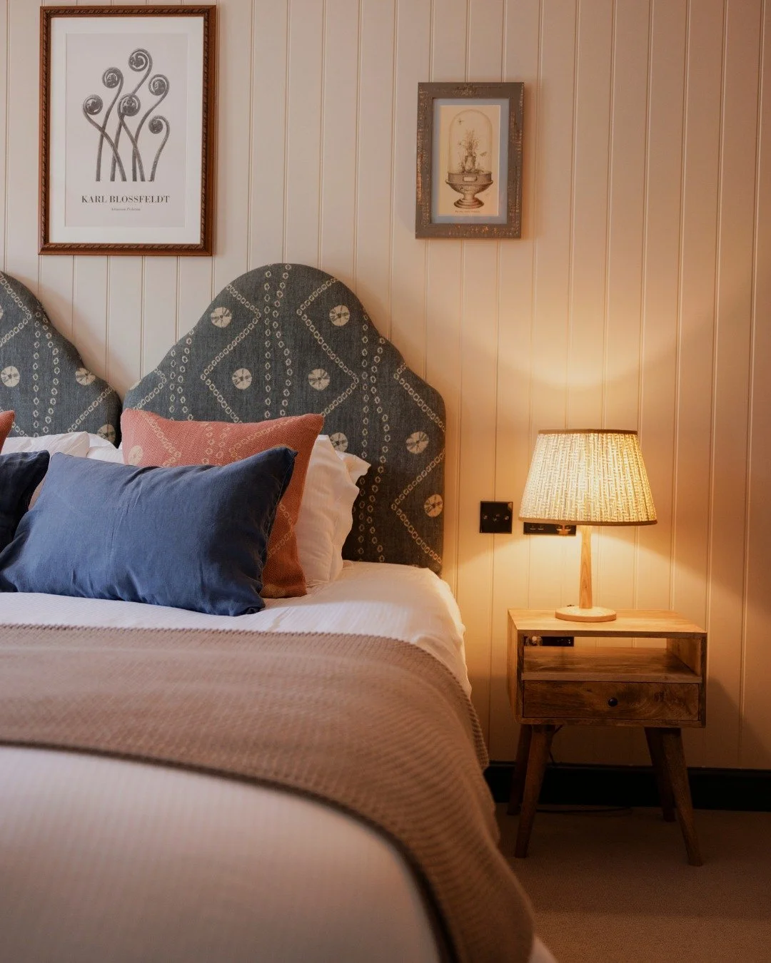 A bedroom corner with a bed, decorative pillows, a wooden nightstand, a lamp, and framed artwork on a wooden-paneled wall.