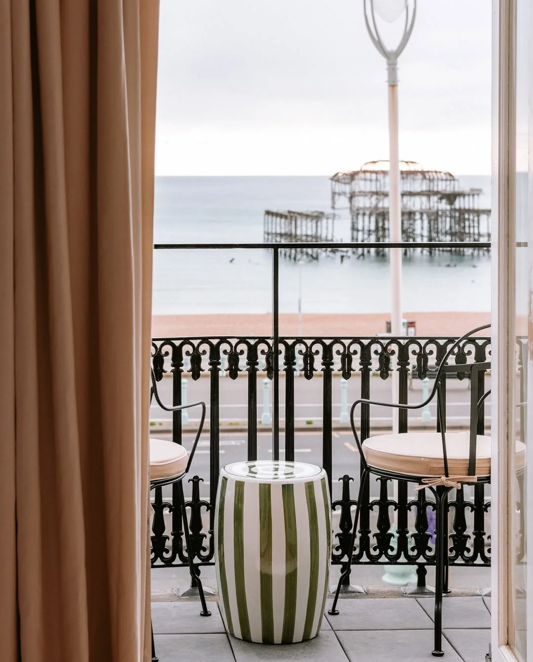 Balcony with two black metal chairs with white cushions, a striped green and white ceramic stool, and a view of the ocean with a pier in the background.