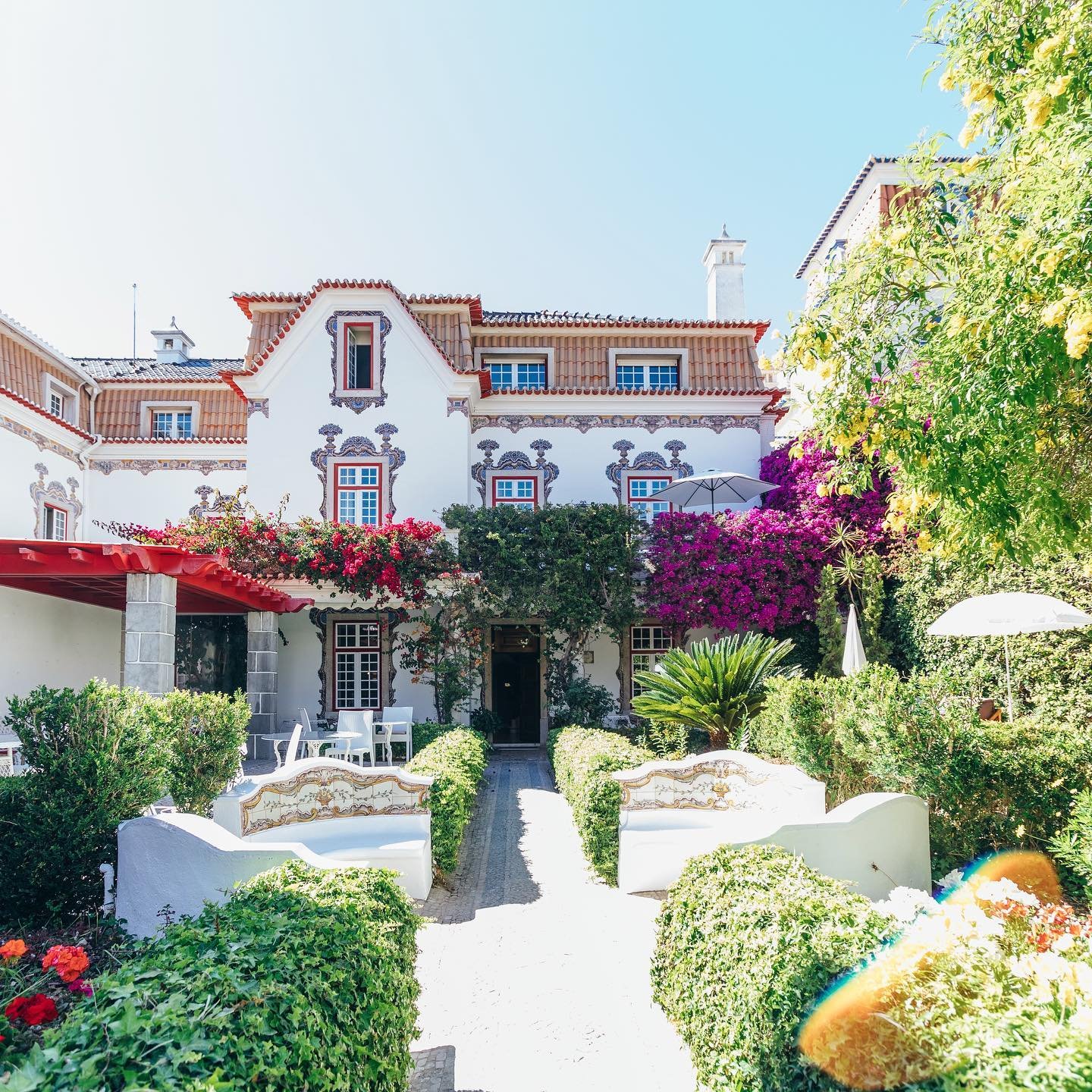A large European-style house with white walls, decorative window trims, red-tiled roof, surrounded by lush garden filled with flowering plants, umbrellas, and outdoor seating.