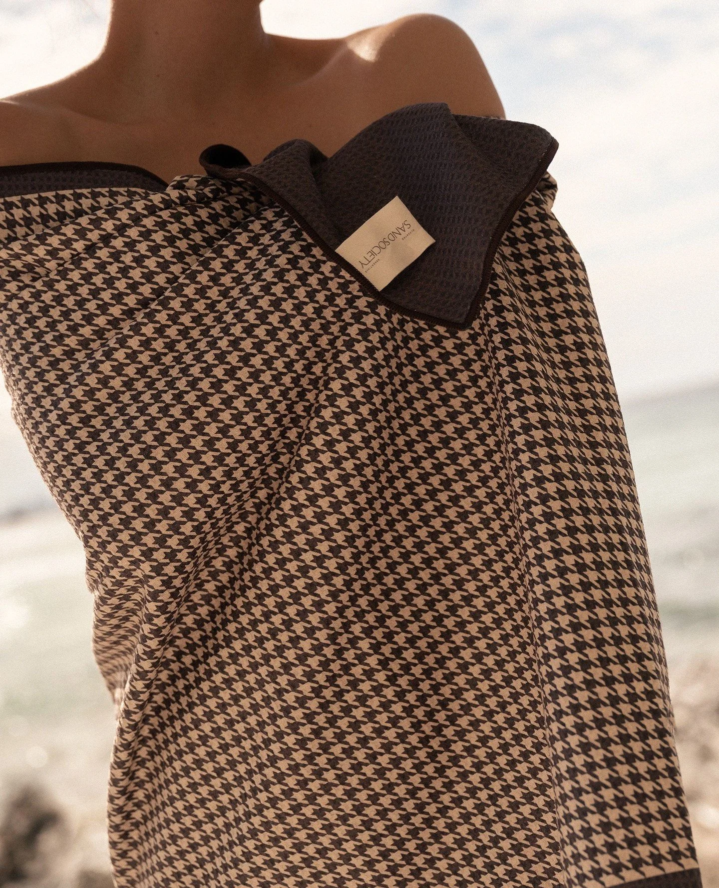 Close-up of a woman wearing a houndstooth patterned shirt and holding a black cloth, with the sky and water in the background.