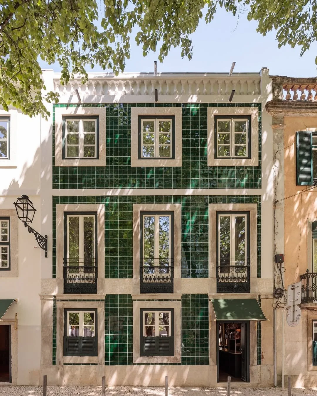 A building with a green tiled facade, three windows with white frames, black railings, and an open doorway with a green awning, beneath leafy trees.
