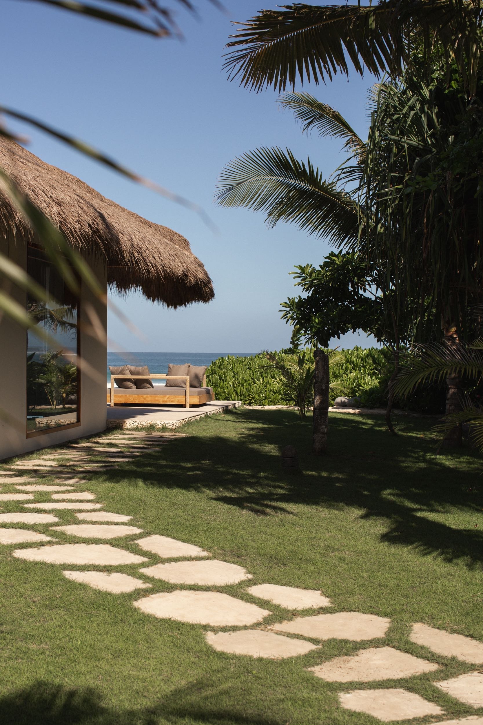 A tropical outdoor setting with a thatched roof structure, a seating area with cushions, lush greenery, palm trees, and the ocean in the background under a clear blue sky.
