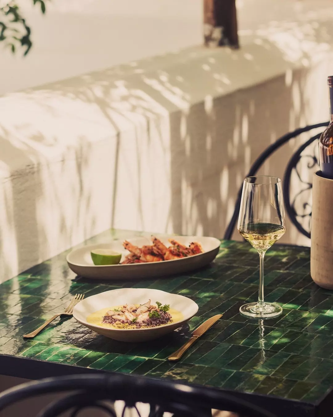 A table with a glass of white wine, a plate of seafood, and another plate of grilled shrimp with a lime wedge. The table has a dark green, marble-like surface and is set outdoors with sunlight casting shadows.