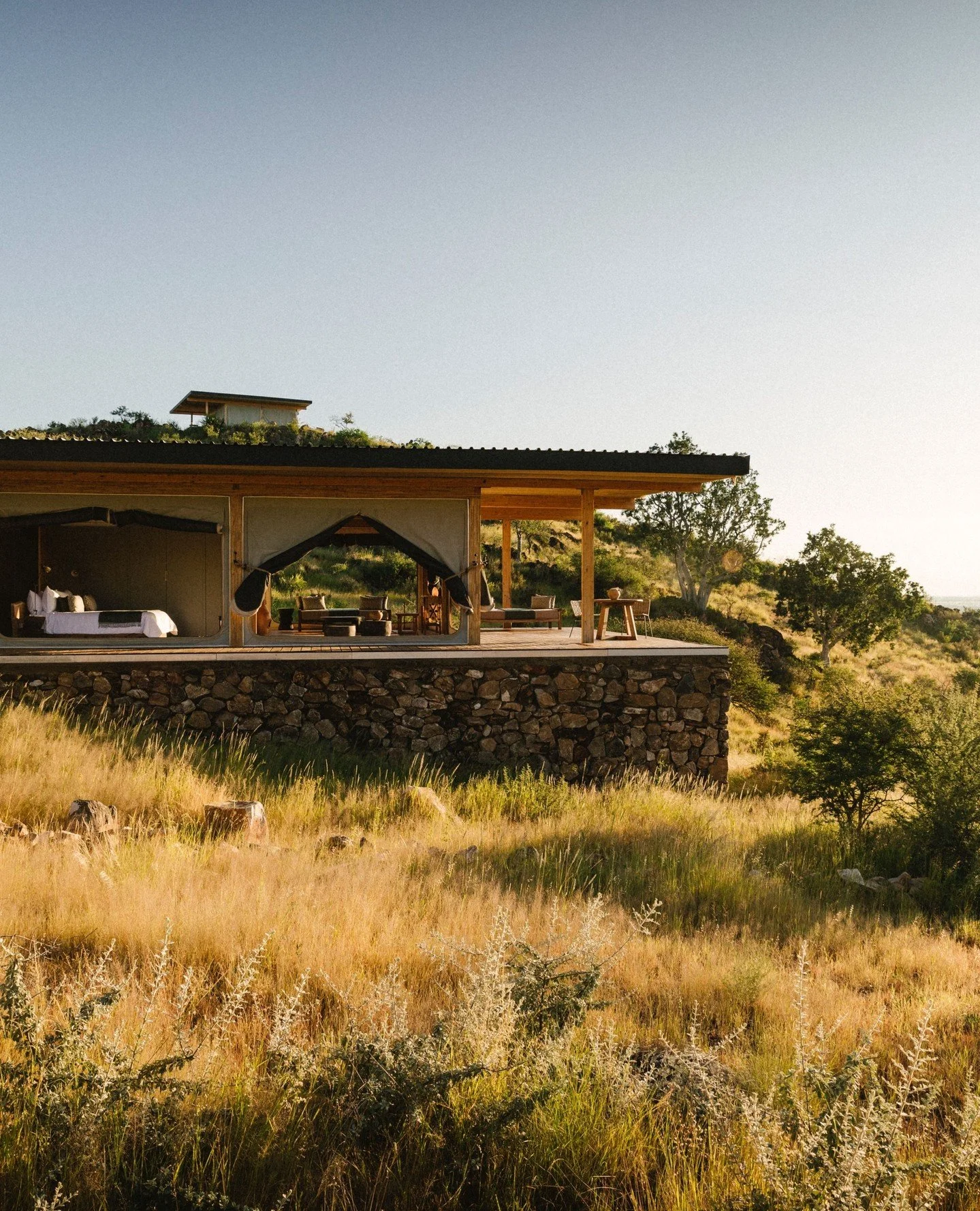 Modern house on a stone foundation with a large outdoor deck, set in a grassy field with trees and a clear sky in the background.