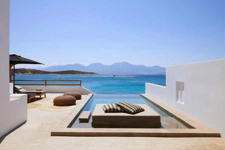 A luxurious outdoor terrace with a view of the ocean, featuring a small infinity pool, striped cushions, patio chairs, and bean bags under an umbrella.