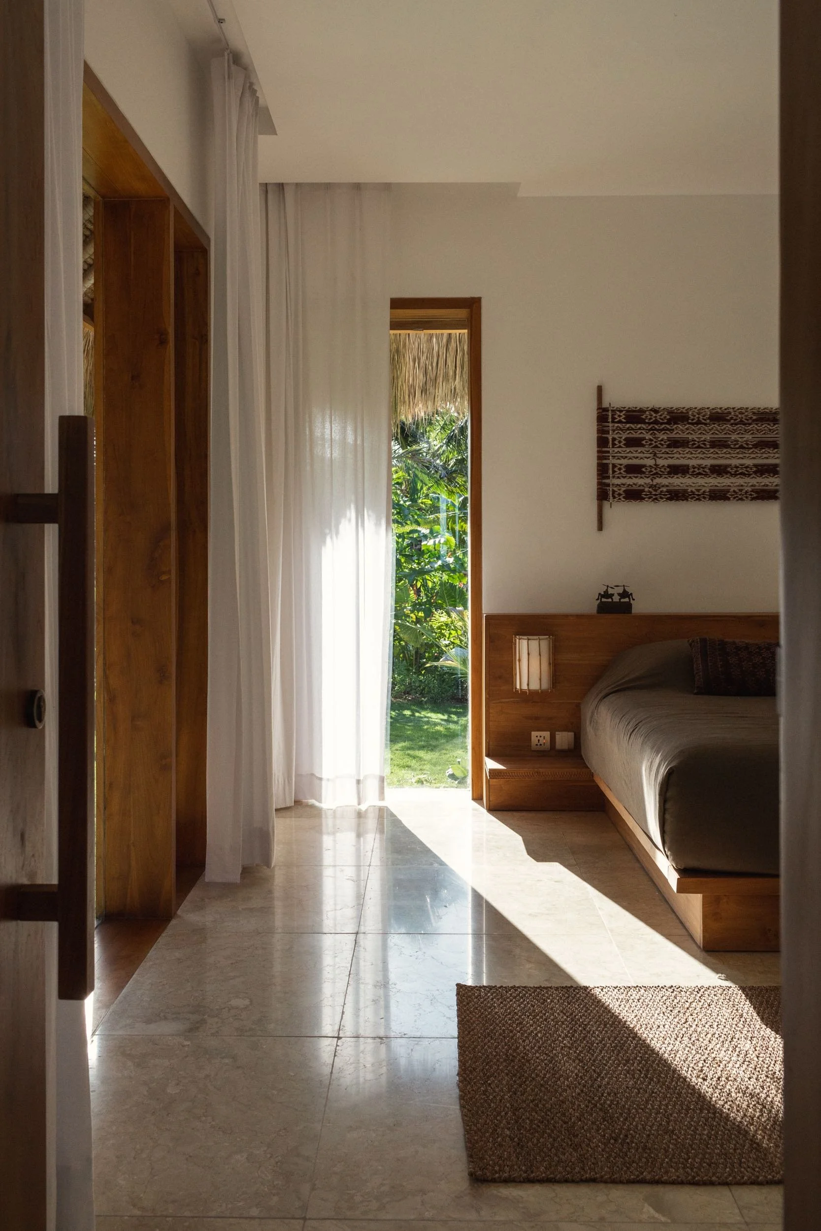 Interior view of a bedroom with a bed, wooden headboard, and a window showing greenery outside. Light streams in through sheer curtains. There's a brown woven rug on the marble floor.
