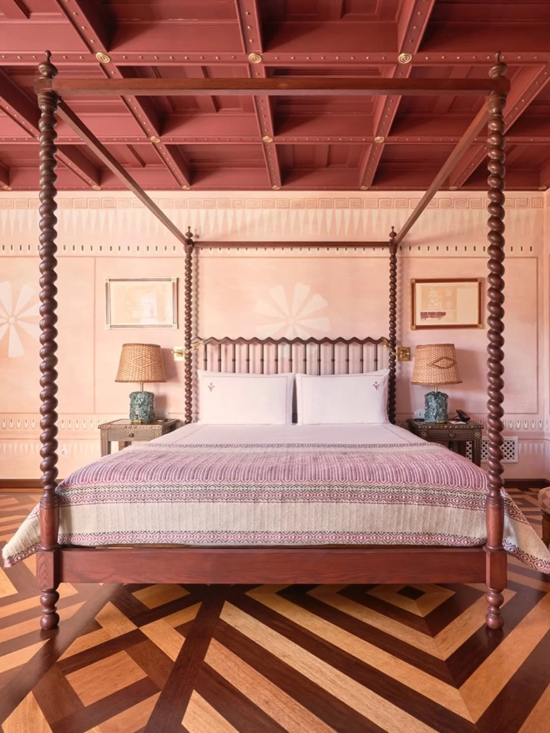 A bedroom with a large four-poster bed with a wooden canopy and twisted posts, pink patterned bedding, two matching nightstands with lamps, and wooden floor with a geometric parquet pattern.