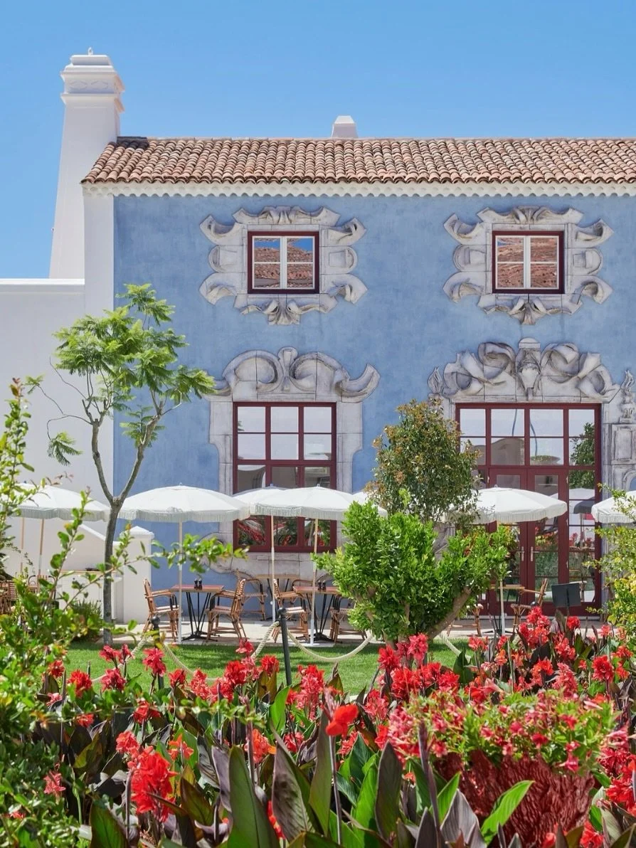 A blue building with decorative window frames, a tiled roof, and outdoor seating with white umbrellas and blooming flowers in the foreground.