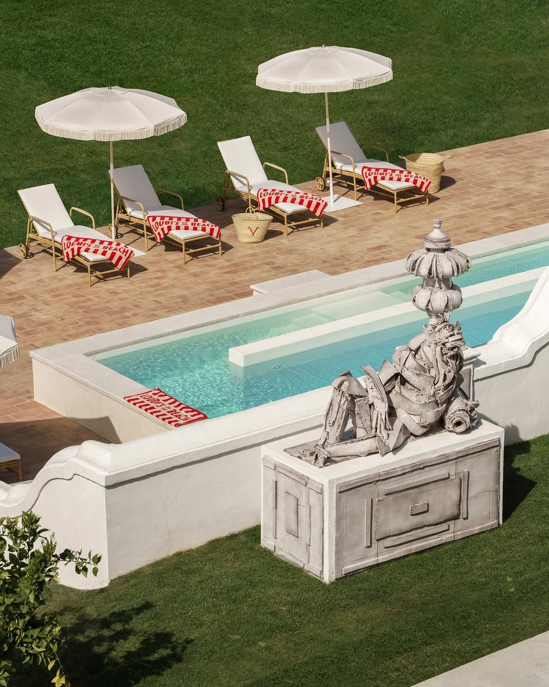 Sun loungers with striped red and white towels, large white umbrellas, and a pool with a fountain, all on a patio with green grass surrounding it.