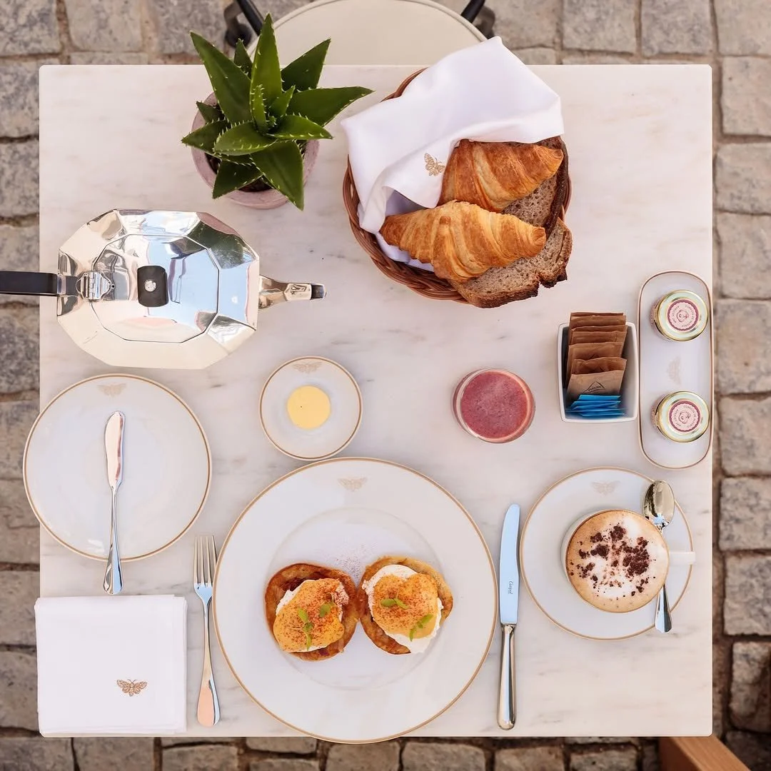 Top-down view of a breakfast table with croissants, bread, a cappuccino, and a glass of juice, complemented by a potted plant and a sugar set.