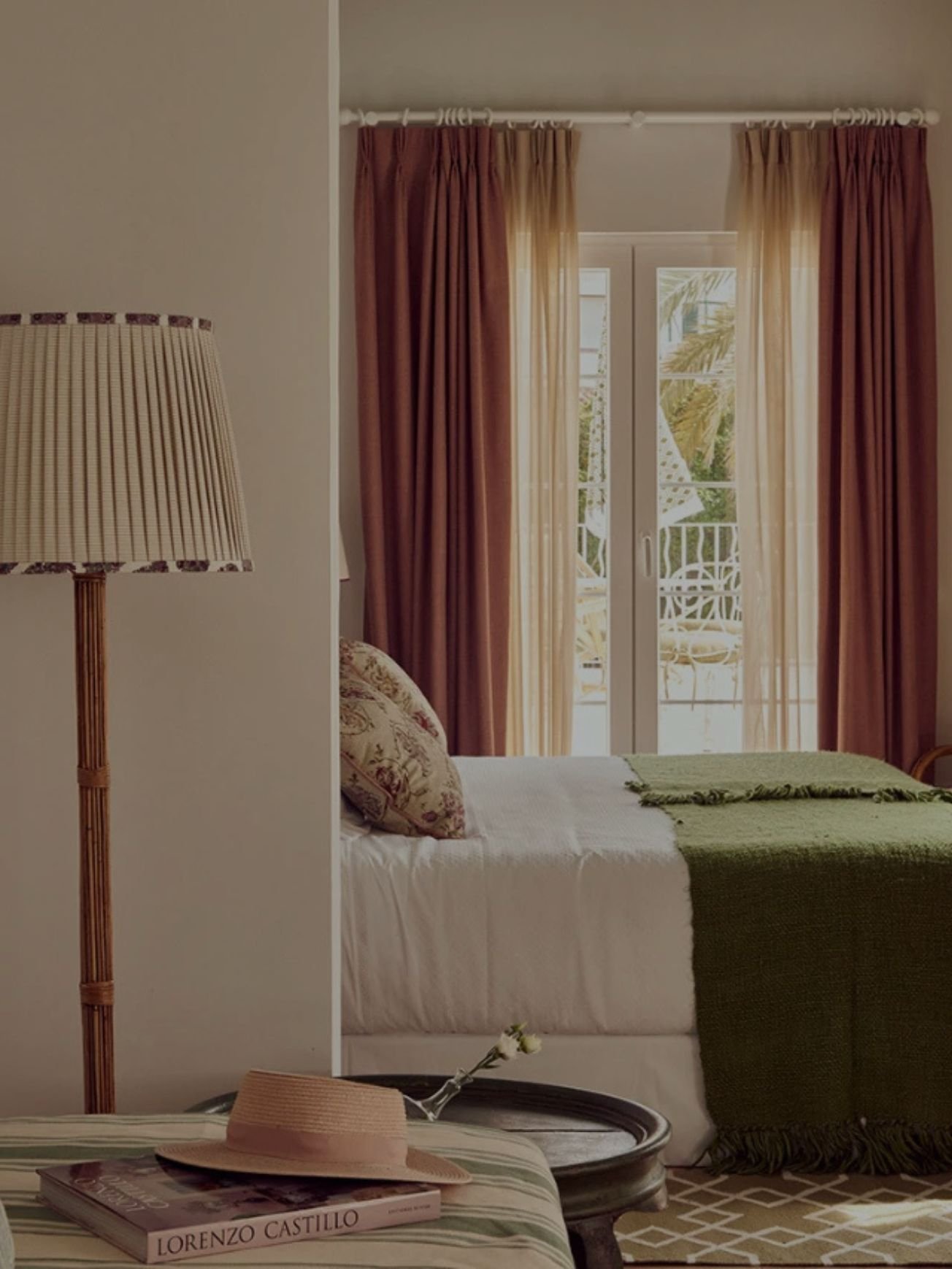 A bedroom with beige walls and large glass doors with curtains leading to a balcony. A bed with floral pillows and a green blanket is visible, with a straw hat, a book titled "Lorenzo Castillo," and a single white flower resting on a small table in the foreground. A tall, striped floor lamp is positioned near the bed.