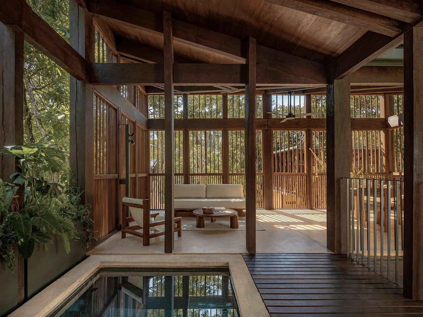 Indoor space with wooden beams, a white sofa, a wooden coffee table, and chairs, surrounded by a railing and trees visible through vertical wooden slats.