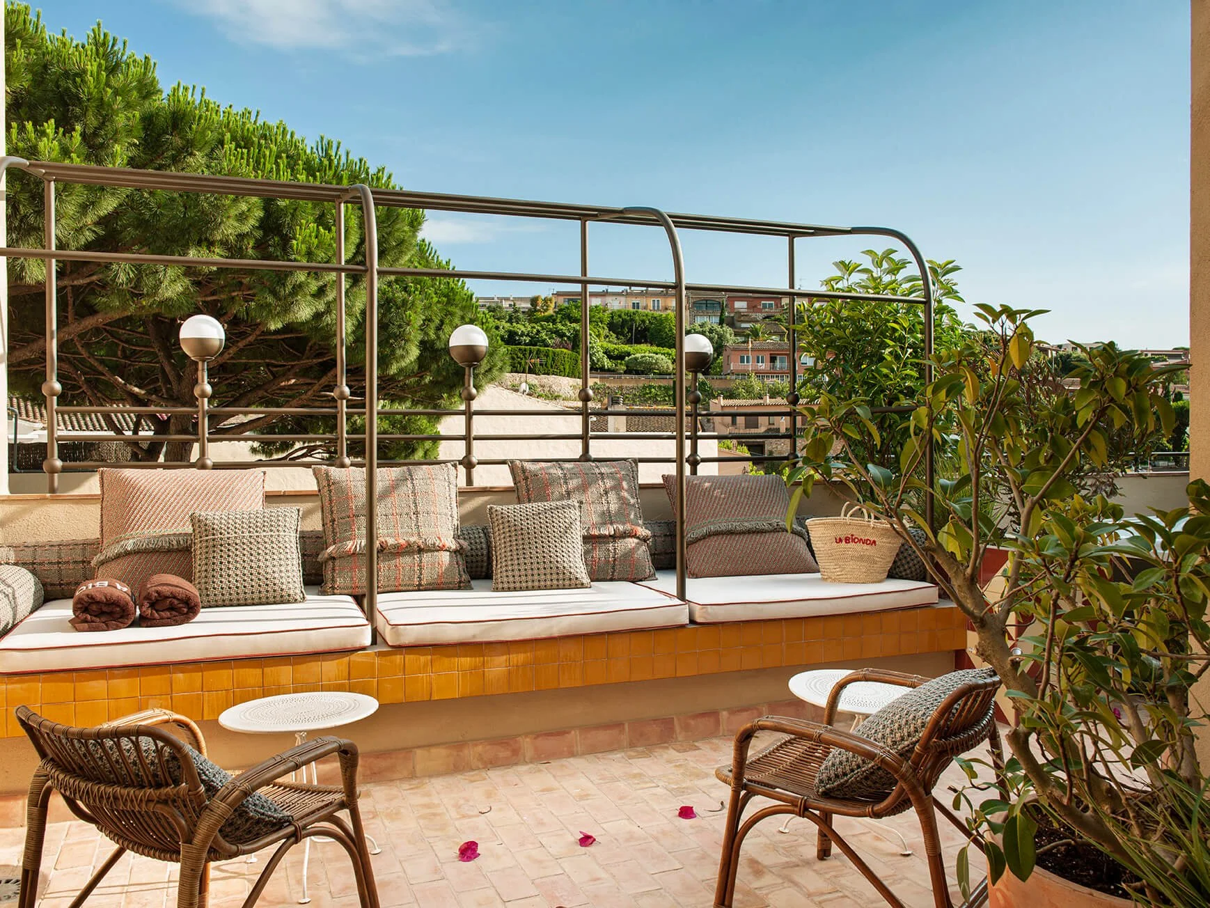 A rooftop terrace with a bench adorned with various patterned cushions, some rolled towels, and a woven bag. A small round white table is near wicker chairs, and cheek pink petals are scattered on the tiled floor. There are lush trees and a view of buildings and greenery in the background, with a partly cloudy sky overhead.