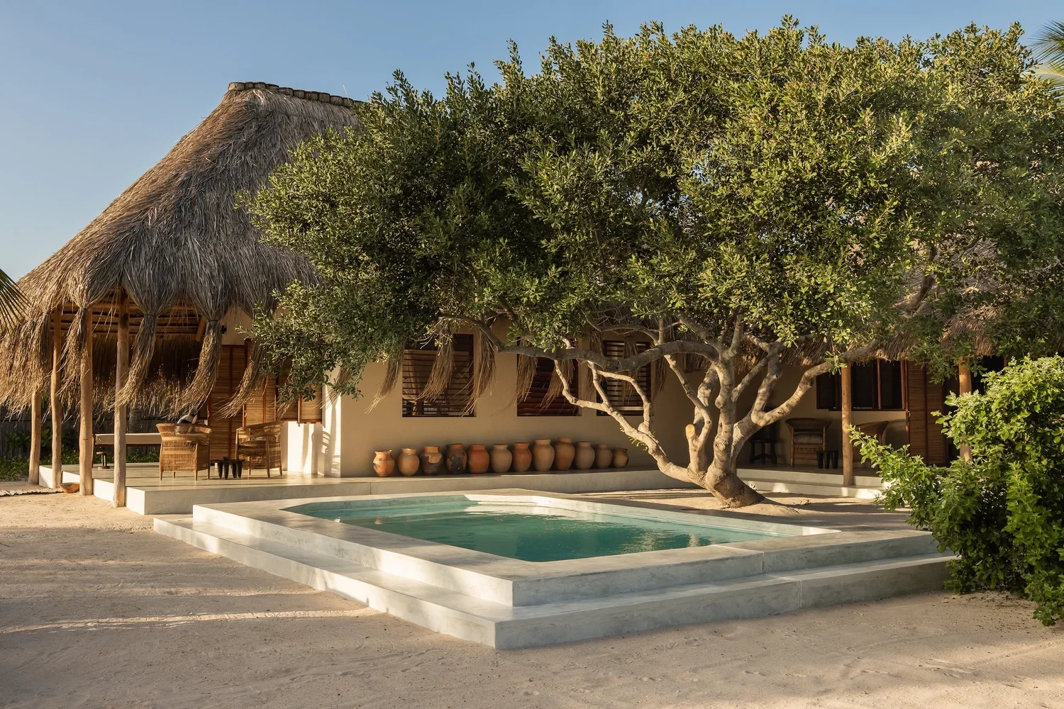 Tropical bungalow with thatched roof, patio area, swimming pool, and large tree providing shade.