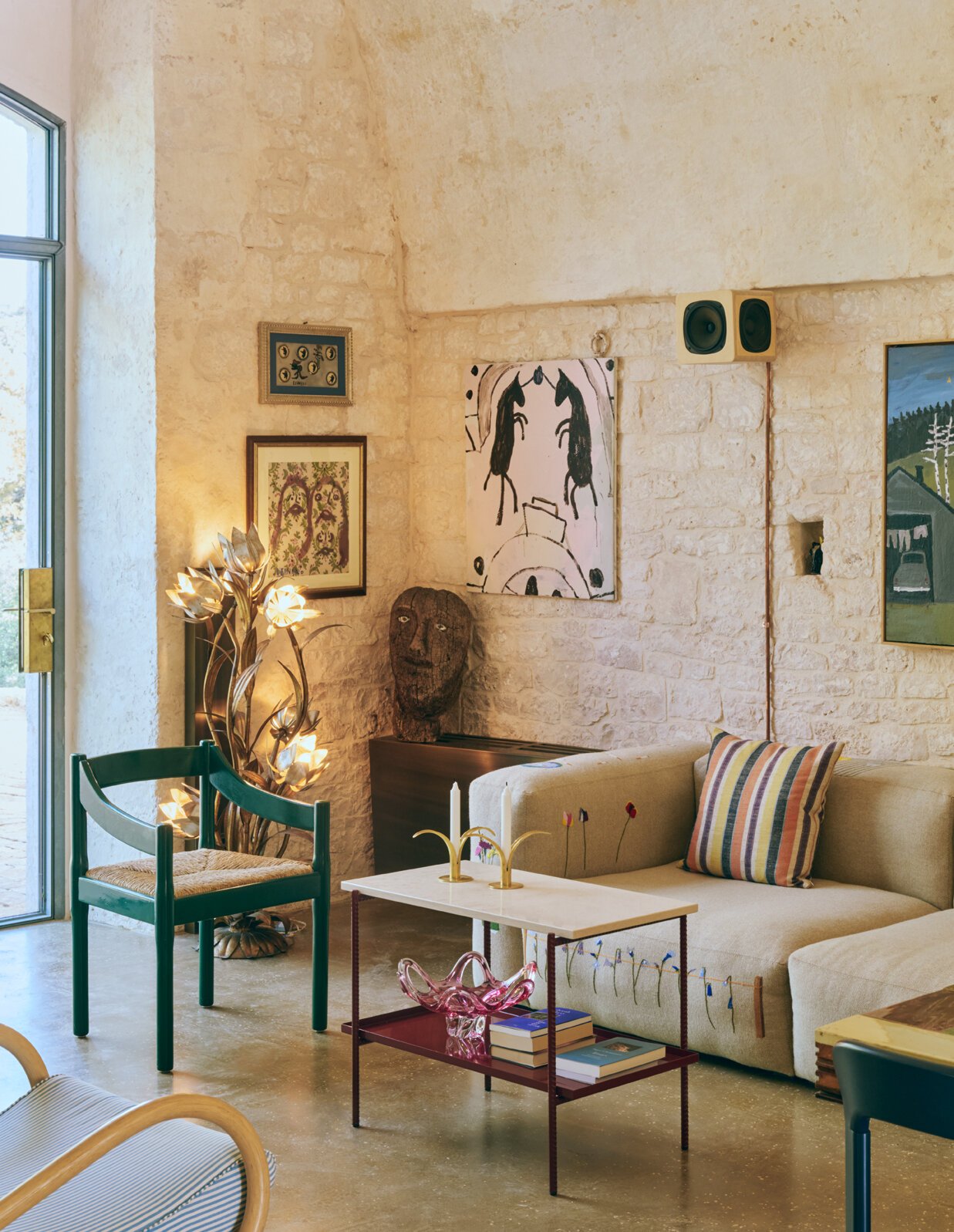 Living room with cream-colored sofa, colorful striped pillow, glass coffee table with art decorations, green chair with woven seat, eclectic wall art, and a large window with natural light.