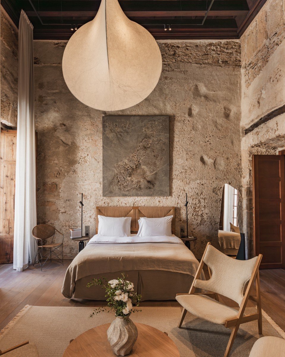 A cozy bedroom with a rustic stone wall, a large bed with a wooden headboard, white pillows, and beige bedding. There are two black bedside lamps, a round wooden table with a vase of white flowers, a beige armchair, a full-length mirror, and a wooden wardrobe. A large, round, paper lantern hangs from the ceiling, and soft natural light filters in from a window with white curtains.