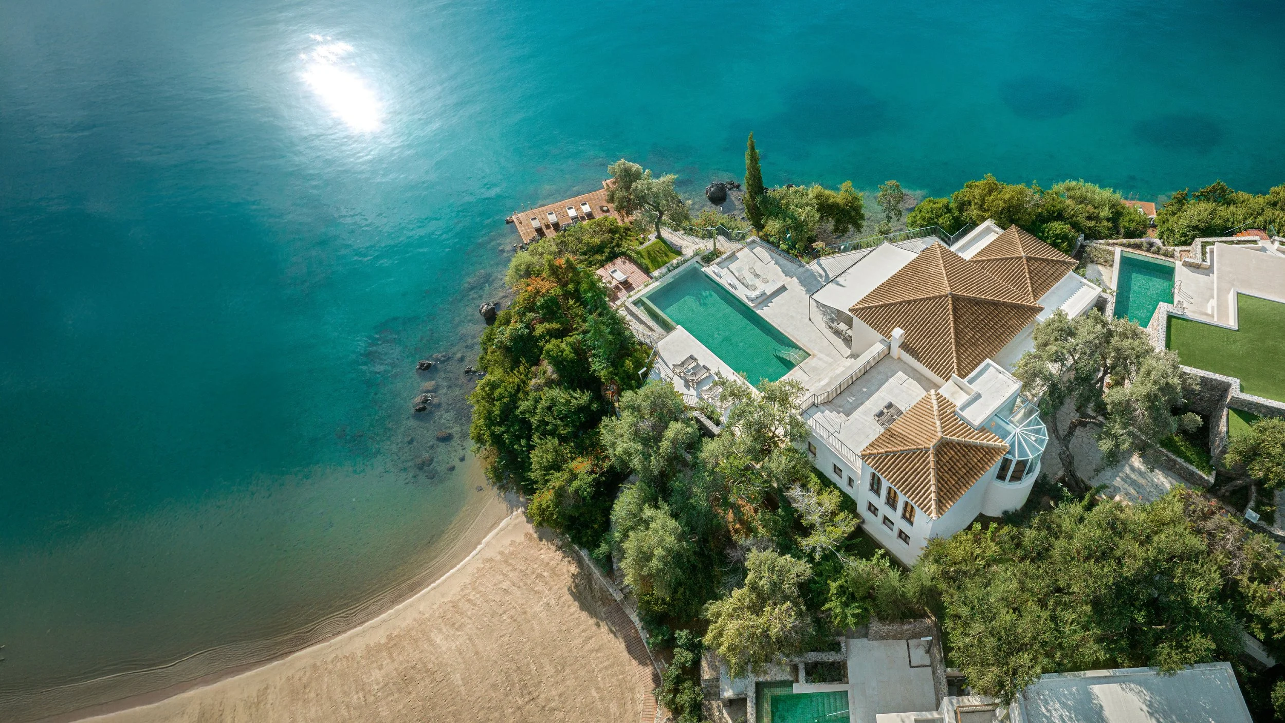Aerial view of a luxurious waterfront property with multiple pools, surrounded by lush greenery, overlooking a calm lake or ocean.