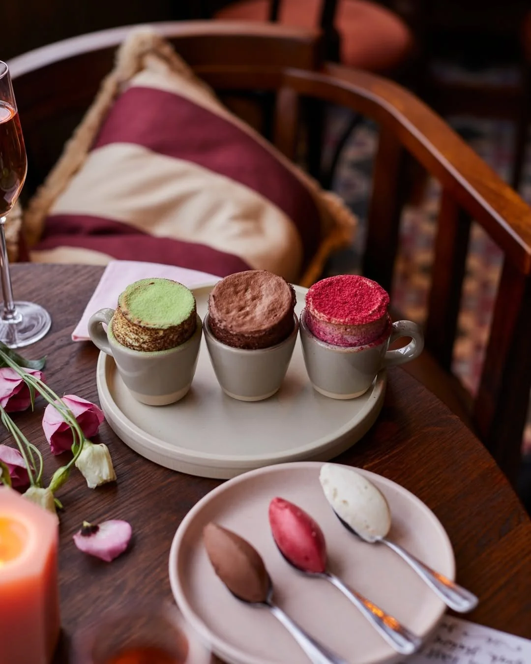 Three cups of hot chocolate topped with colorful powders, served on a tray on a wooden table, with spoons of different flavors on a side plate, in a cozy setting with flowers and a glass of wine nearby.