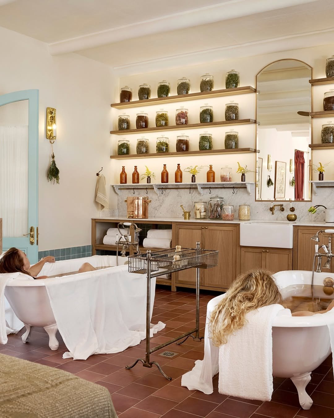 Two women relaxing in vintage bathtubs in a cozy, well-decorated bathroom with shelves of jars filled with herbs and flowers, a large mirror, and warm lighting.